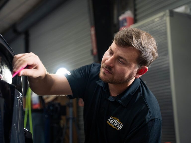 Mechanic inspecting a car panel with a tool in a garage, wearing a black polo shirt