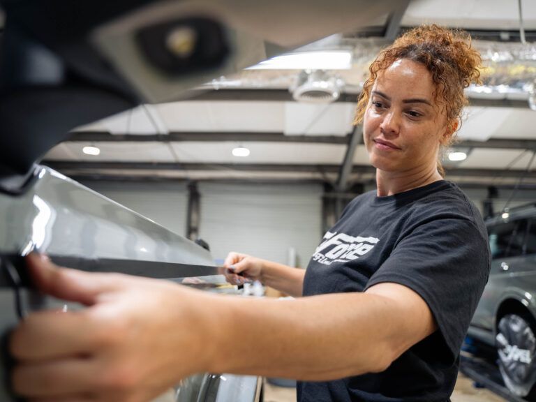 Woman in a garage wiping a car window with a cloth, with a black car in the background.