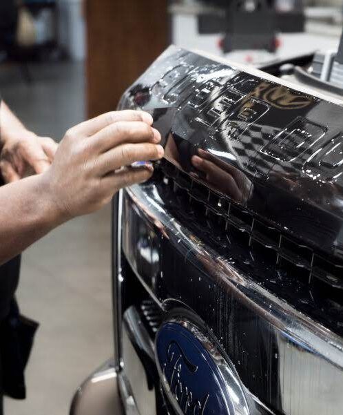 Person detailing a silver car’s front bumper in a garage, seated on a rolling stool