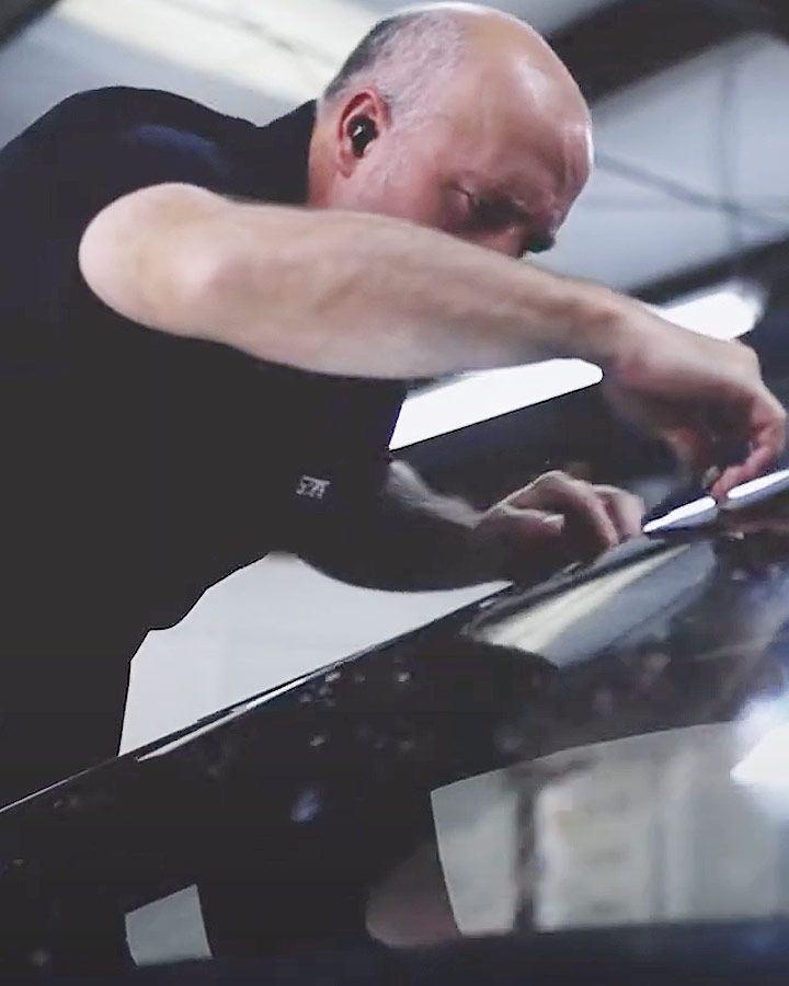 Person in a black shirt leaning over and working on a glossy car windshield indoors