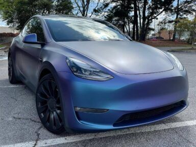 Blue Tesla parked in a lot, front-left view, with black wheels and trees in the background