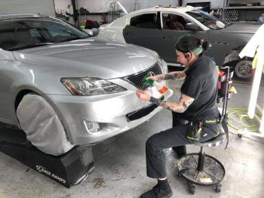 Person detailing a silver car’s front bumper in a garage, seated on a rolling stool