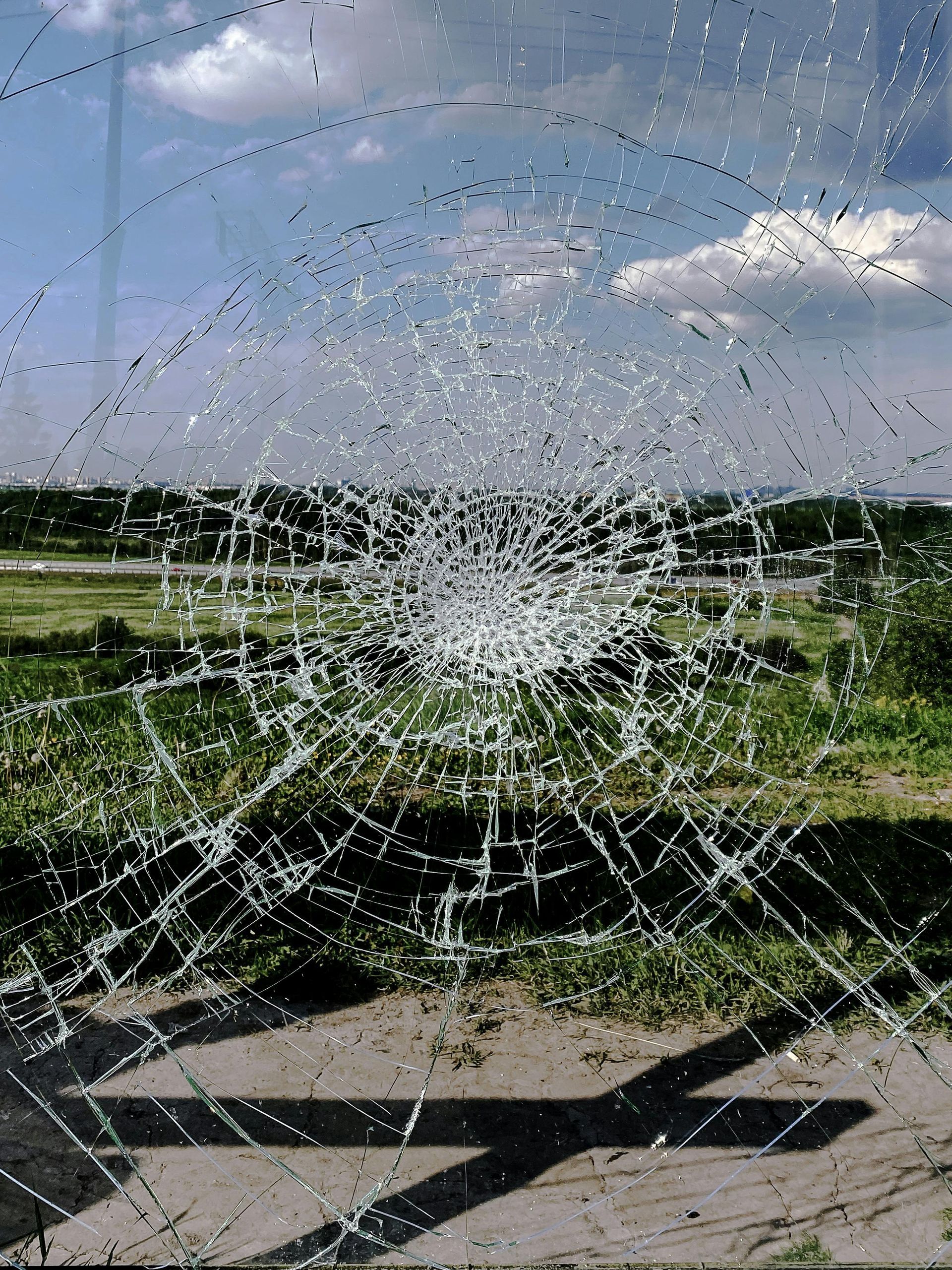 Cracked glass pane with a shattered hole, sky and grassy field visible behind it.