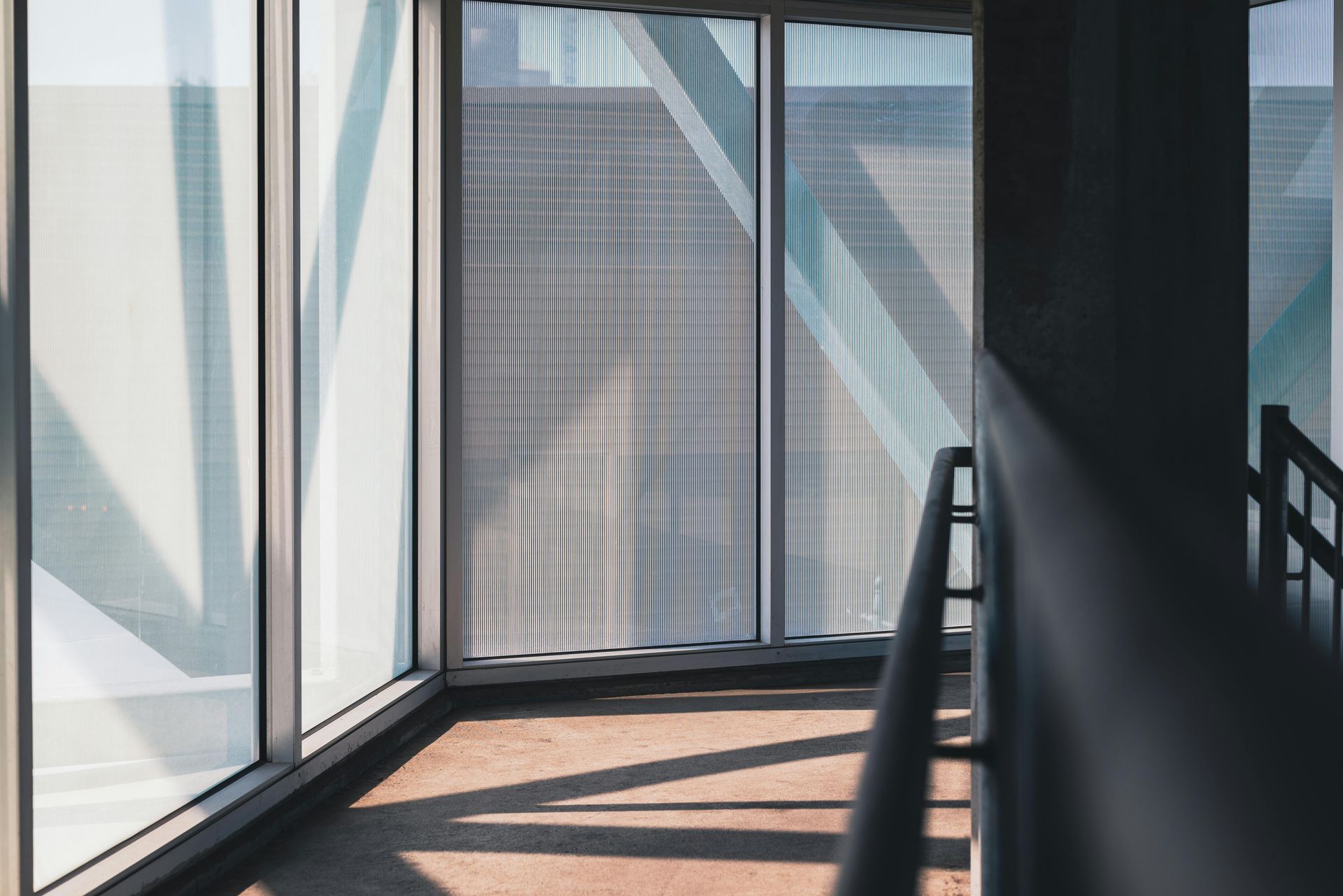 Sunlit walkway with glass walls, metal railings, and diagonal shadows indoors