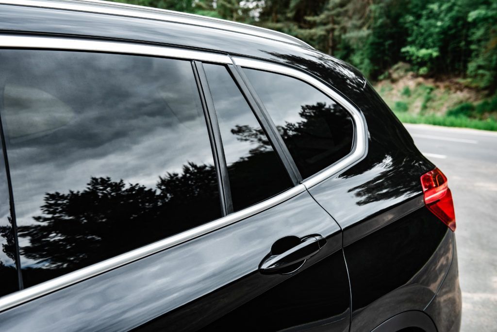 Black car side window and rear door on a road with trees reflected in the tinted glass