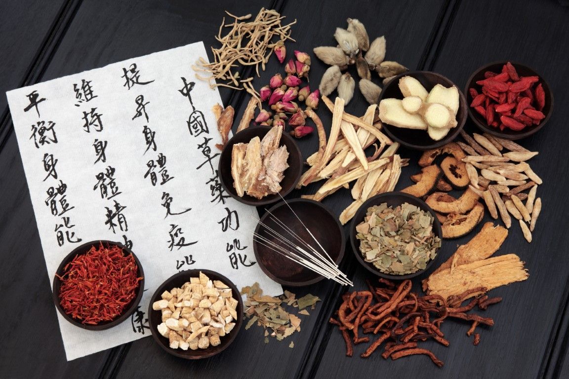 Assorted dried herbs, roots, and berries arranged on a dark surface with Chinese herbal labels