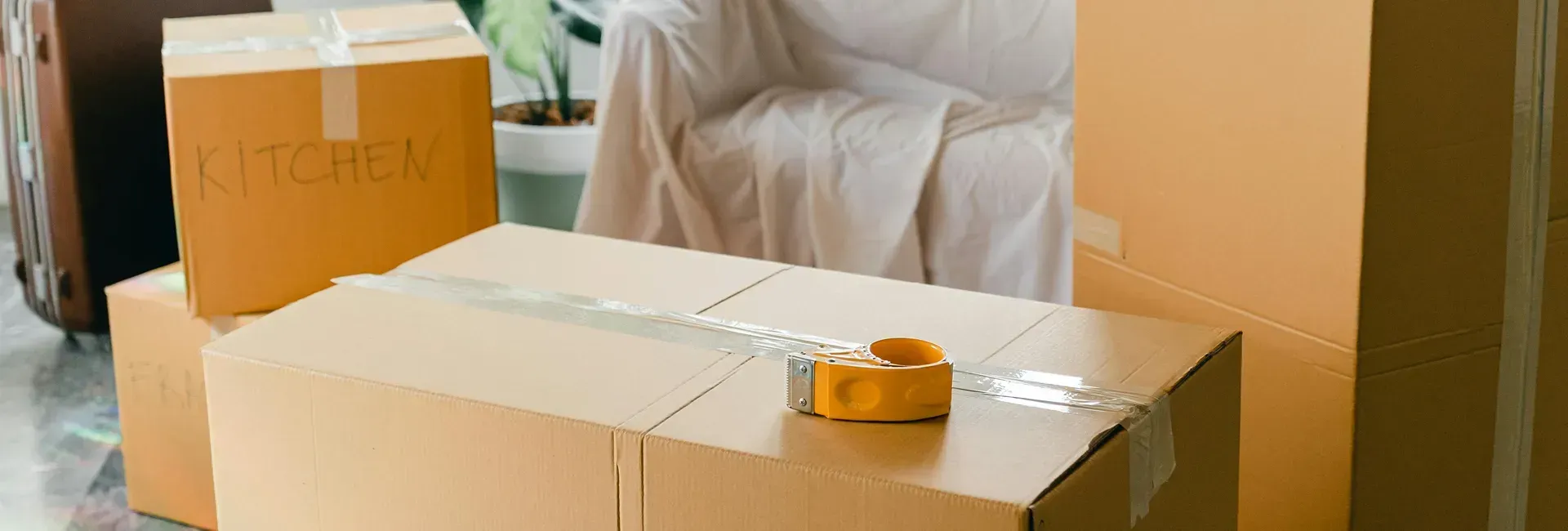 Cardboard boxes stacked for moving, tape dispenser on the largest box, chair covered by a sheet in background.