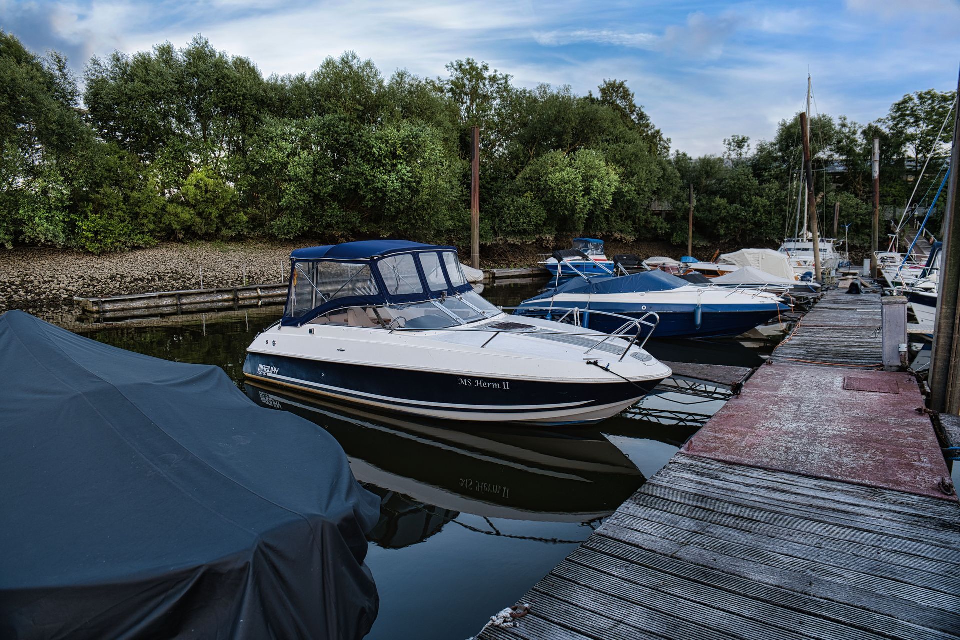 Boats docked along a marina beside a tree-lined canal, with a covered boat in the foreground.