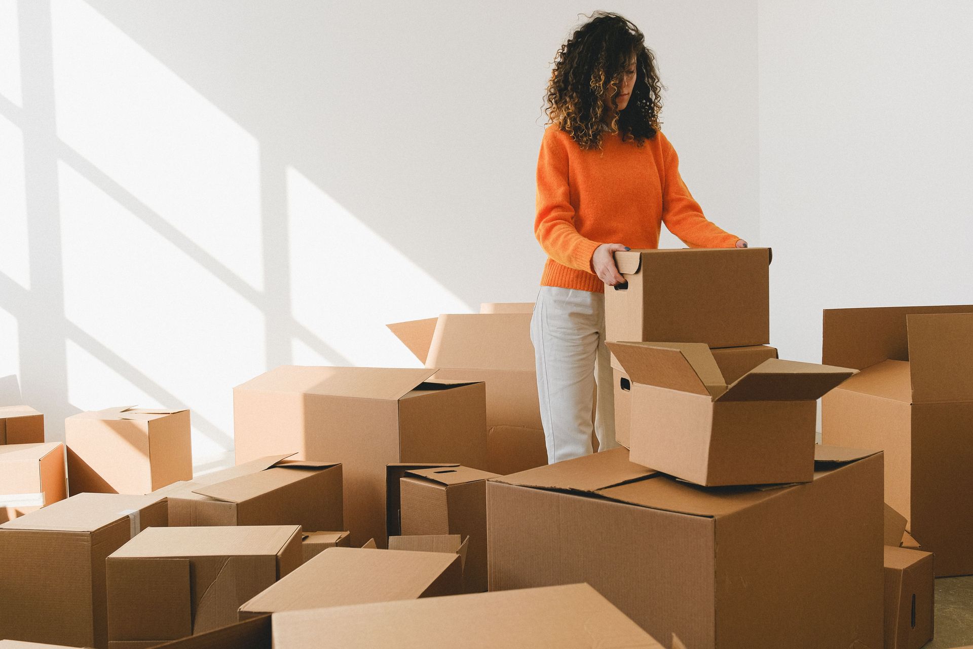 Person unpacking cardboard boxes in a sunlit room with a white wall and window shadows.