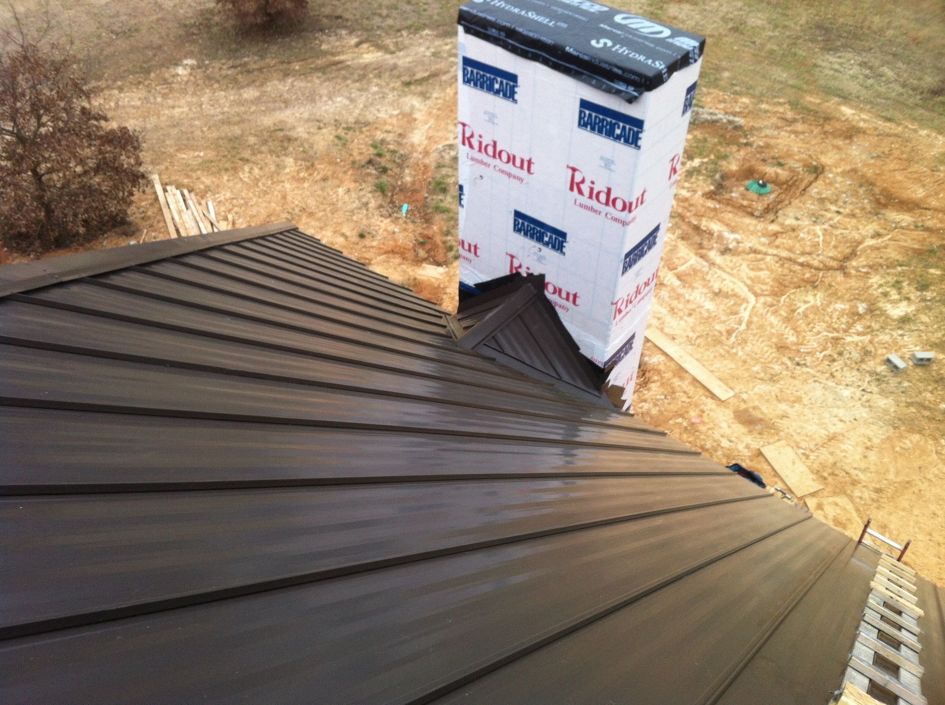 Dark metal roof under construction beside wrapped chimney on a dry, sandy lot