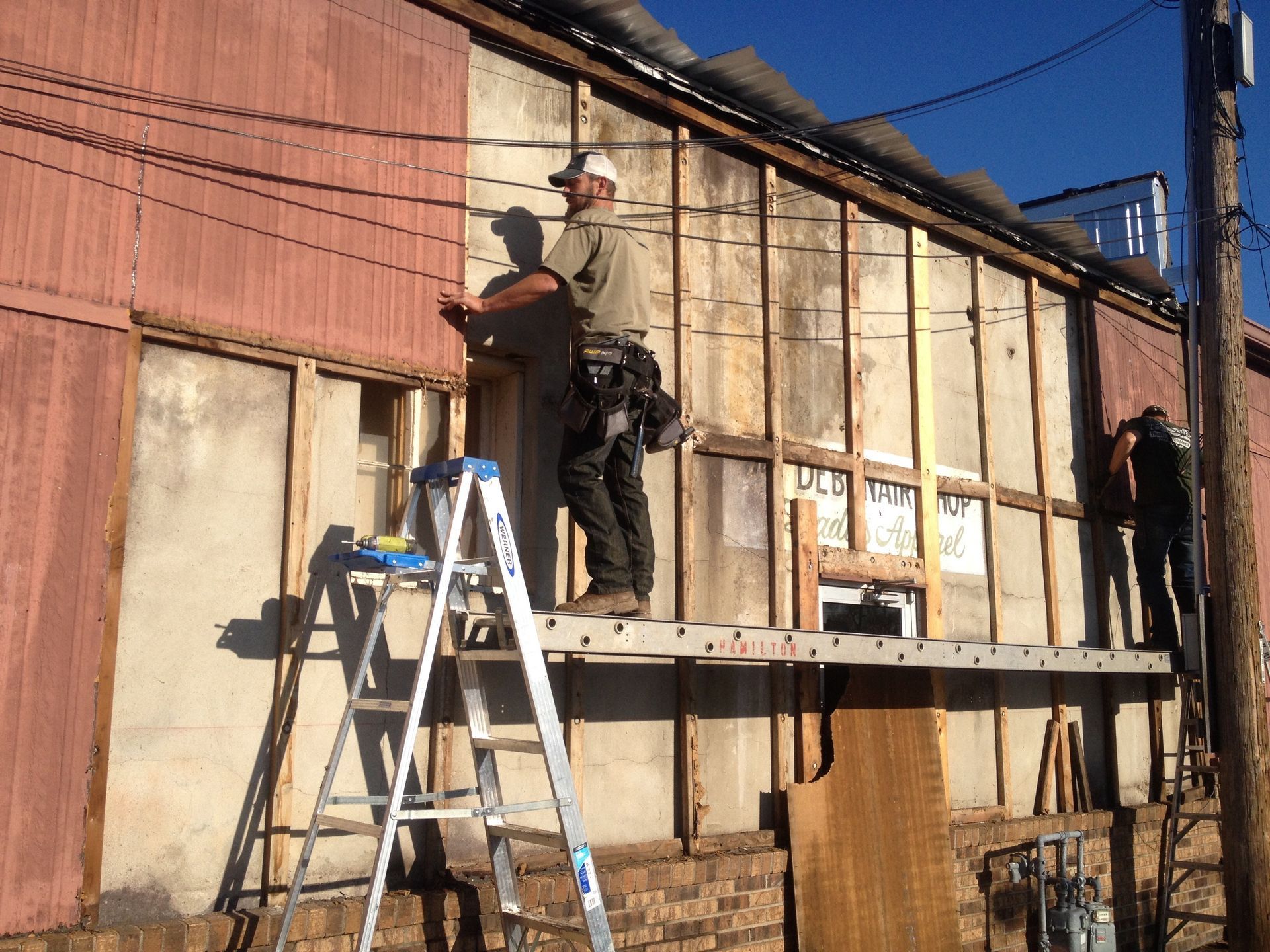 Workers on ladders repairing the side of a weathered building under a clear blue sky.