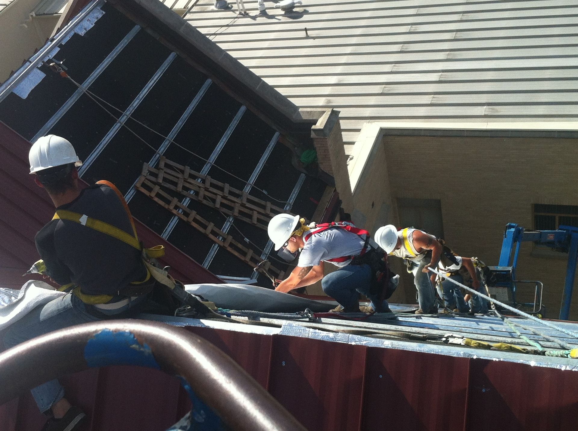 Workers in hard hats and safety gear on a steep roof, repairing damage near a tilted skylight.