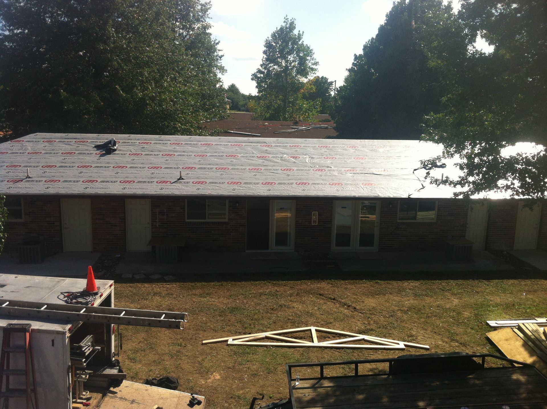Single-story brick house with damaged shingled roof and construction debris in the yard, trees behind.
