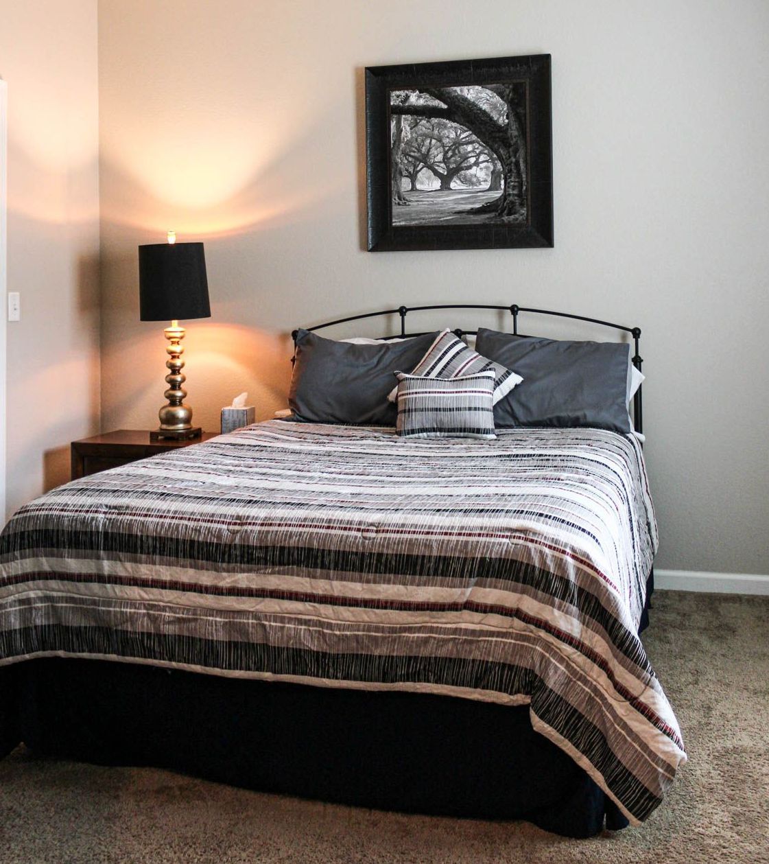 A neatly made bed with a patterned gray and white quilt, dark pillows, a bedside lamp, and framed wall art.
