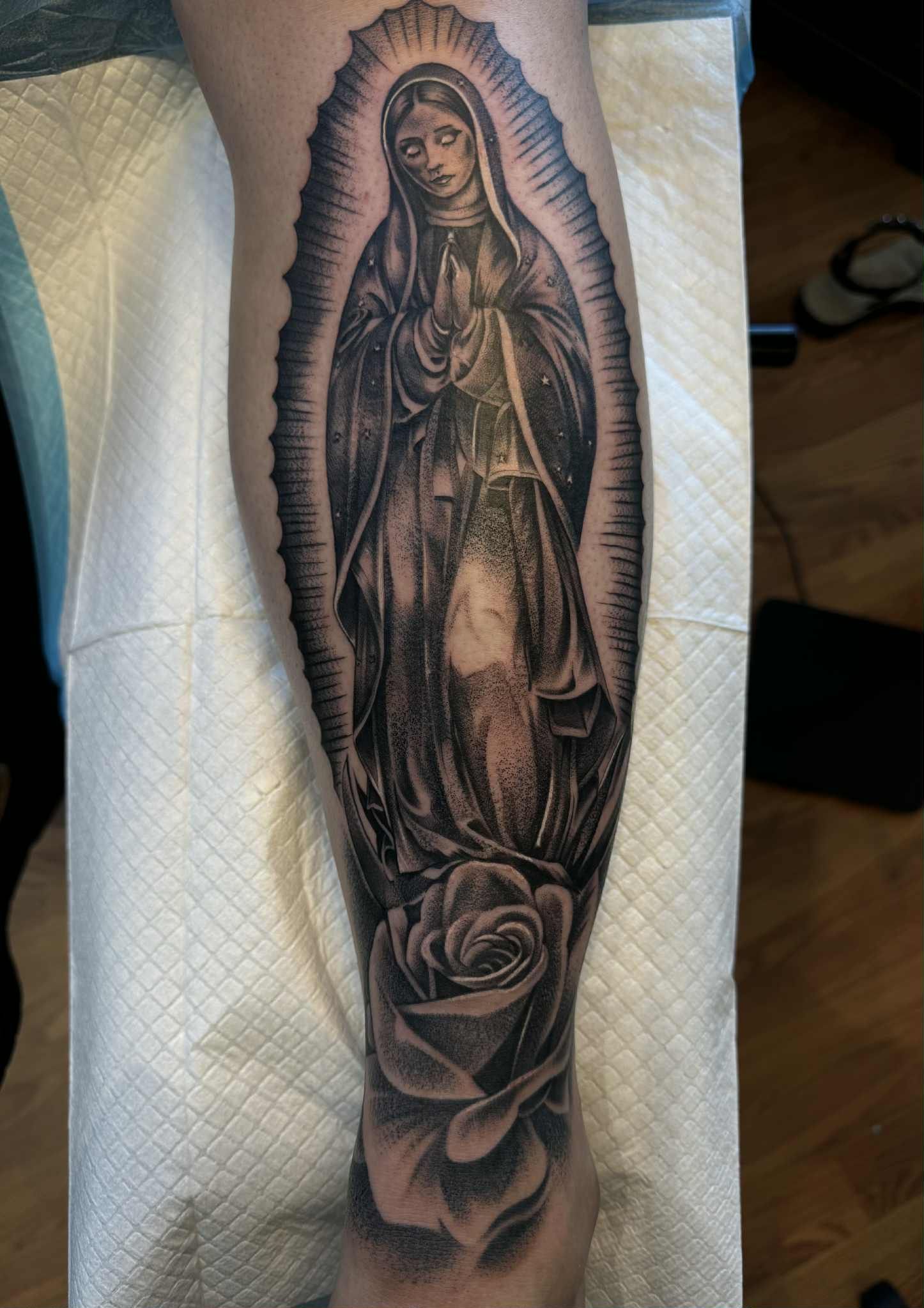 Tattoo of the Virgin Mary inside an oval frame, above a rose on a lower leg.