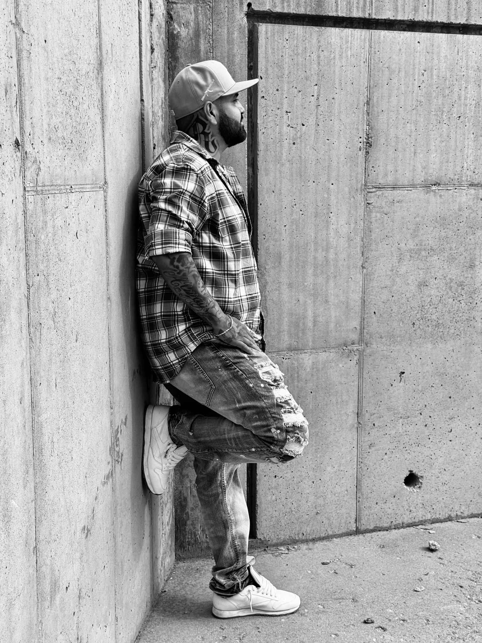 Man in checkered shirt leans against a concrete wall, right leg bent, wearing a cap.
