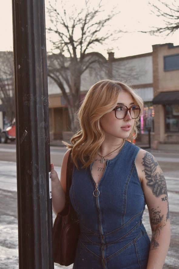 Woman in denim dress and glasses leans on a pole, looking away. Downtown street.