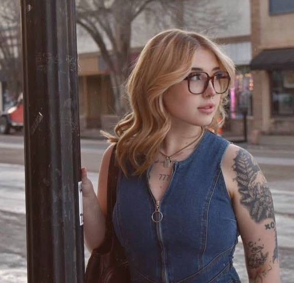 Woman with glasses in denim dress, holding a pole on a city street, looking away.