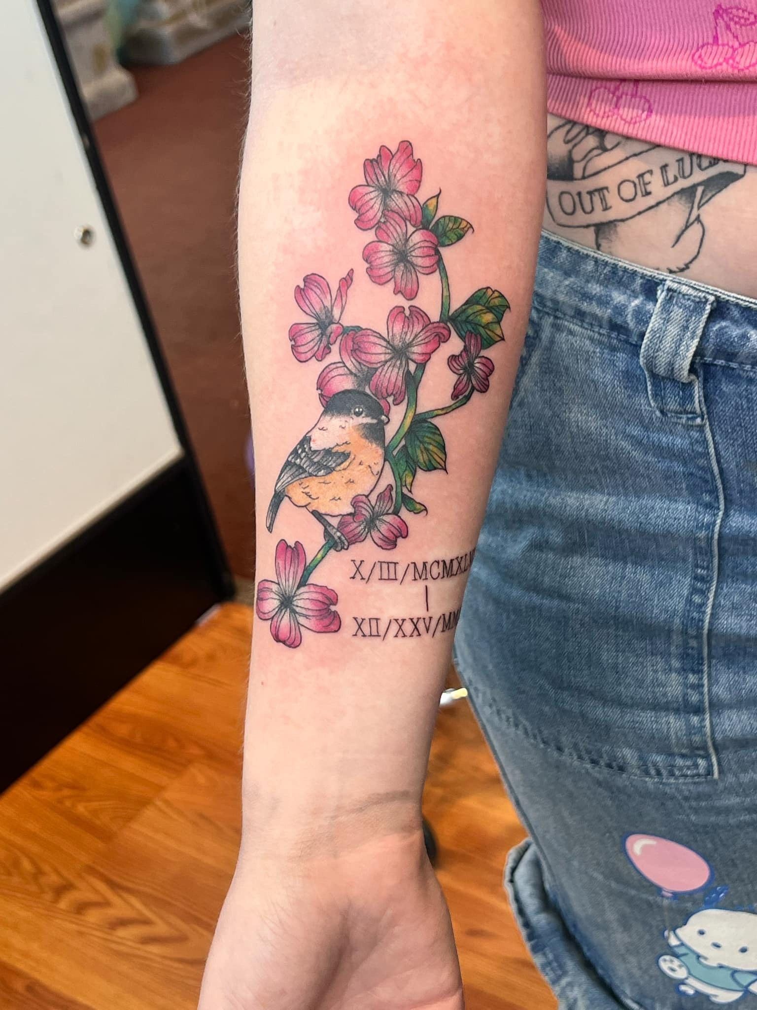 Tattoo of a bird on a flowering branch with text on the forearm. Pink flowers, green leaves.