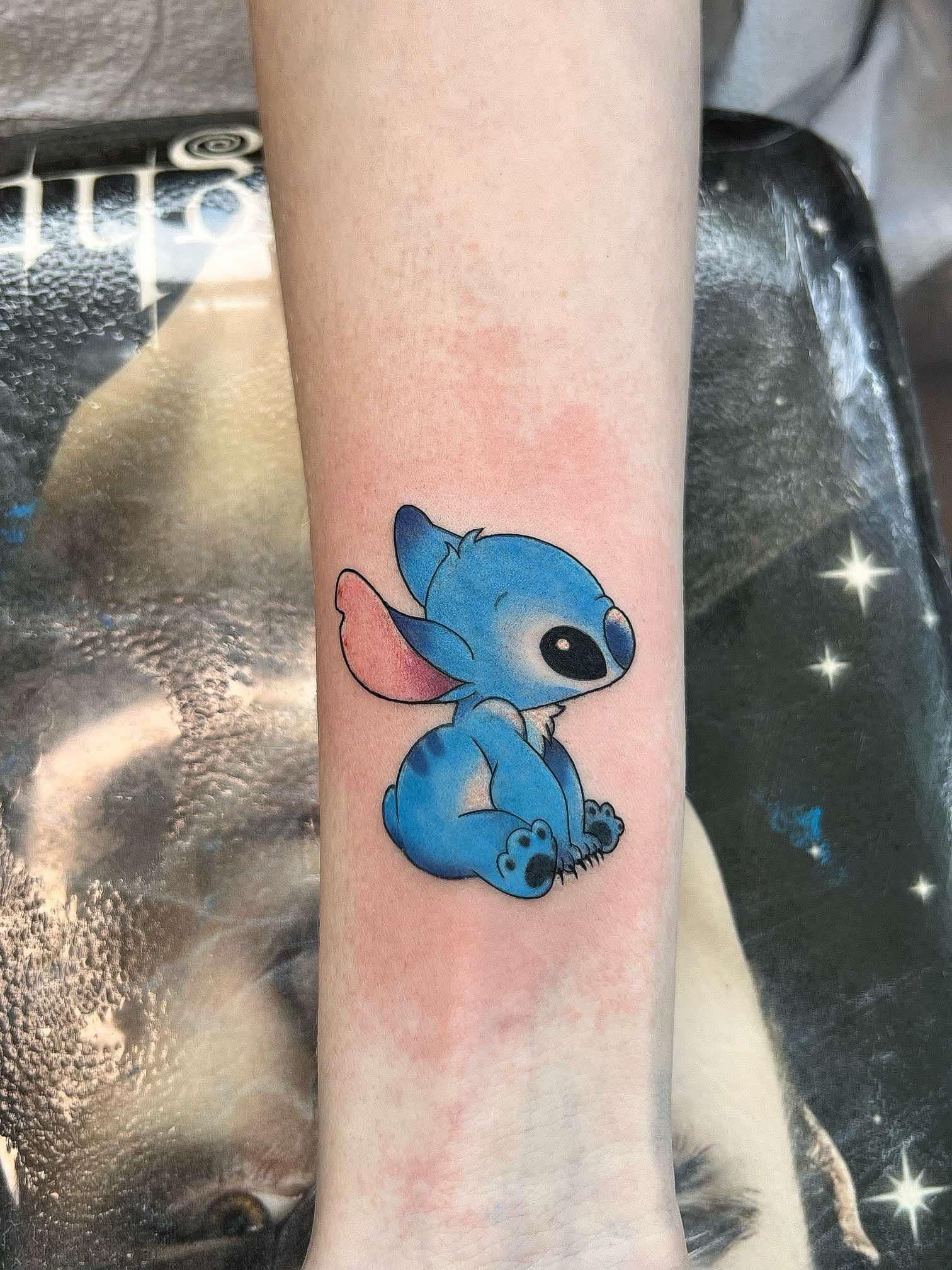Blue Stitch tattoo on a pale arm, seated, with black outline and blue shading.