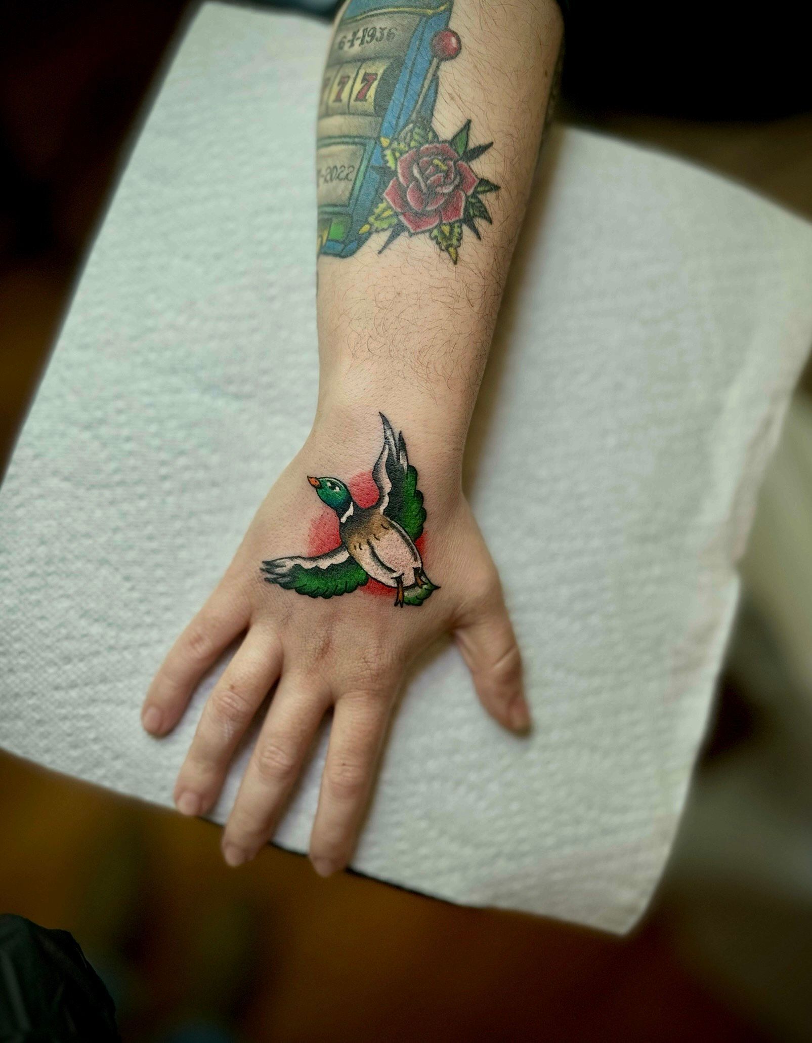 Hand tattoo of a flying duck, green, white, brown, and red, on a person’s hand. Tattooed arm above.