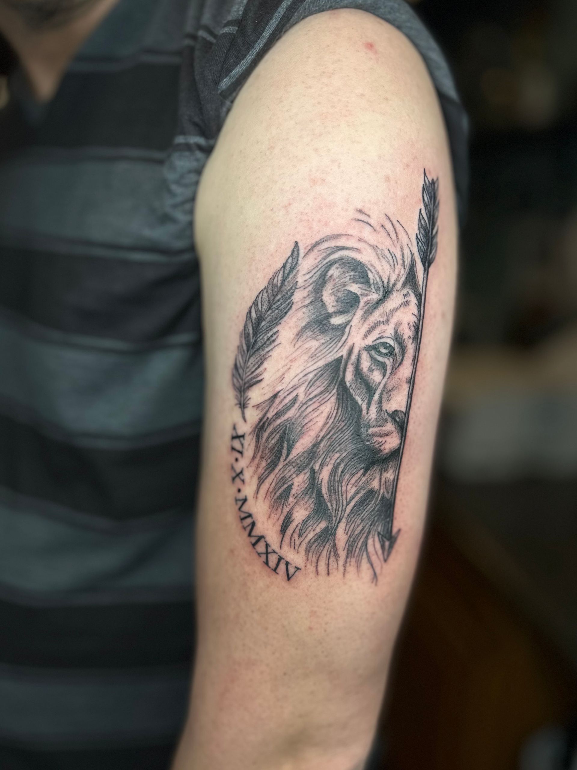Tattoo of a lion's face with feathers, roman numerals, on a person's upper arm.