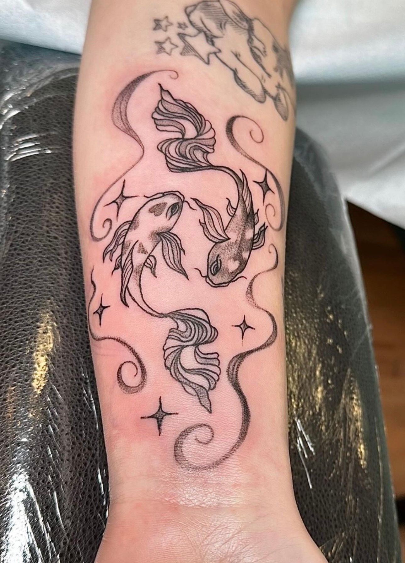 Black line drawing tattoo of two fish, stars, and swirls on a wrist.