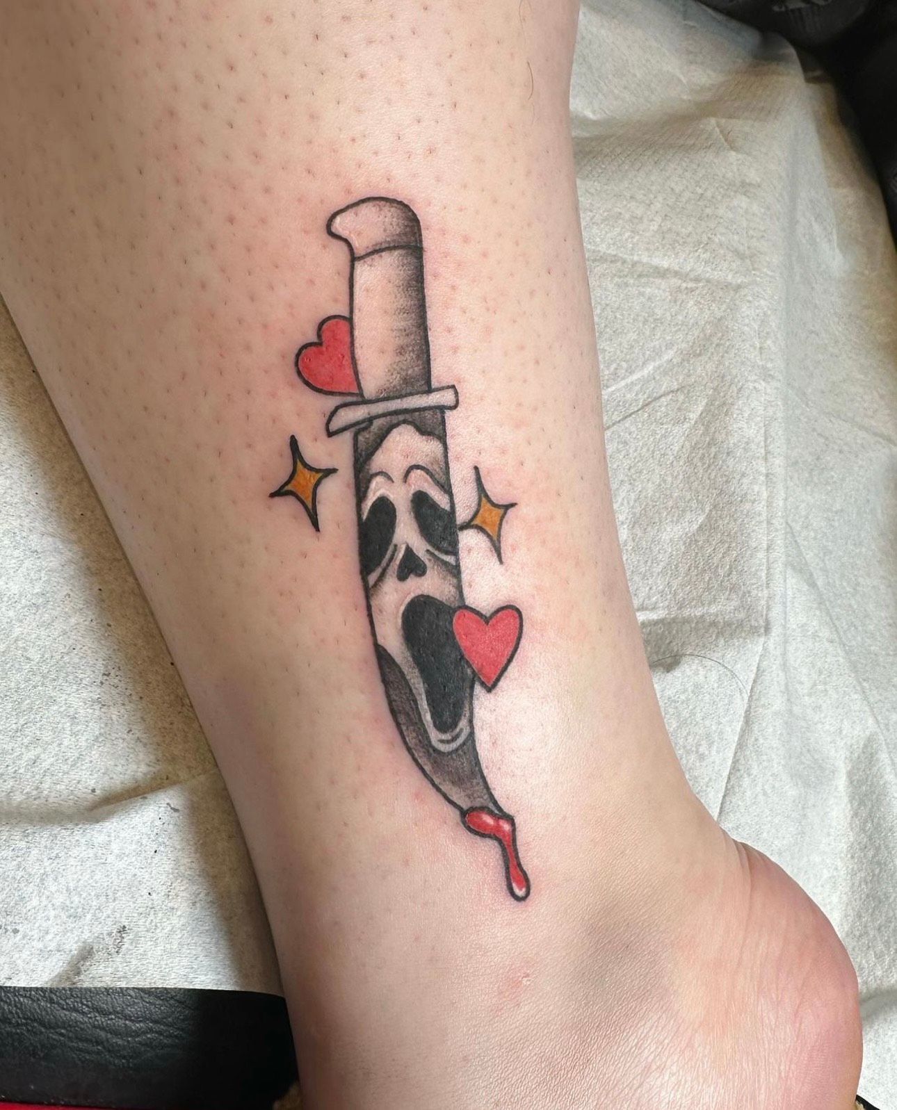 Tattoo on ankle of Ghostface killer knife with hearts and sparkles. Blood drips from blade.