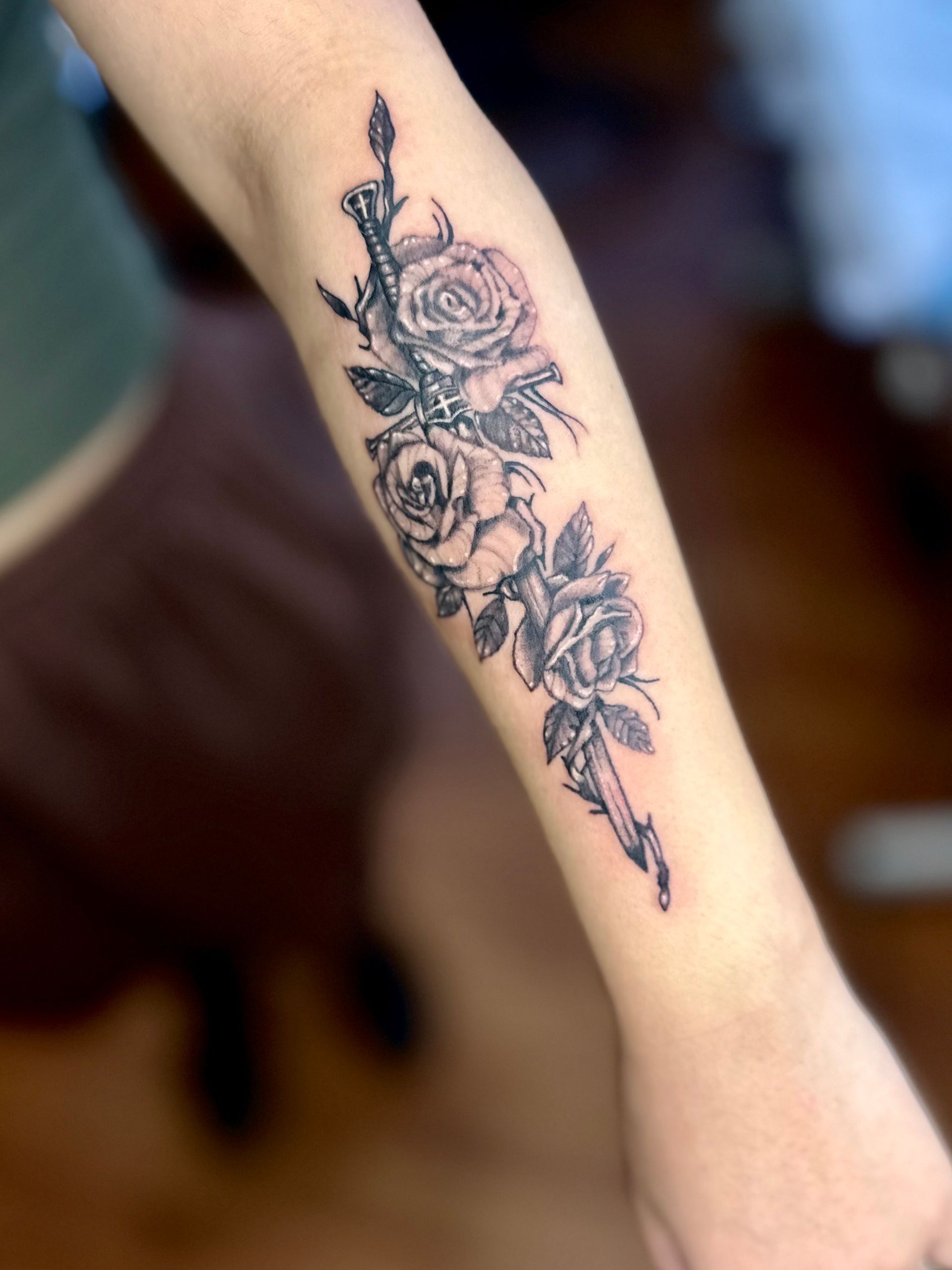 Forearm tattoo: roses entwined with a dagger and a decorative piece, all in black ink.
