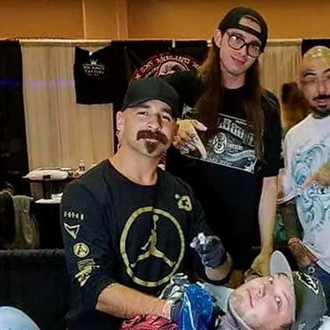 Tattoo artist giving a tattoo at a convention, with two assistants and a partially visible client.