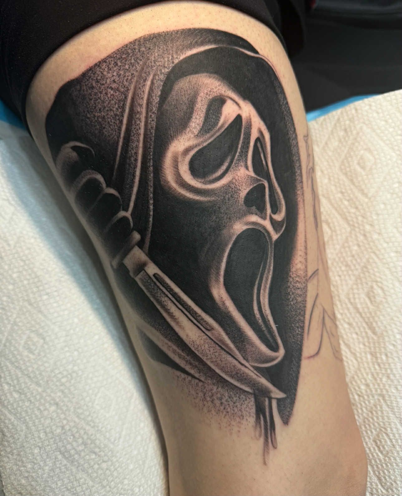 Tattoo of Ghostface mask and knife from the Scream movies, black and white shading on a leg.