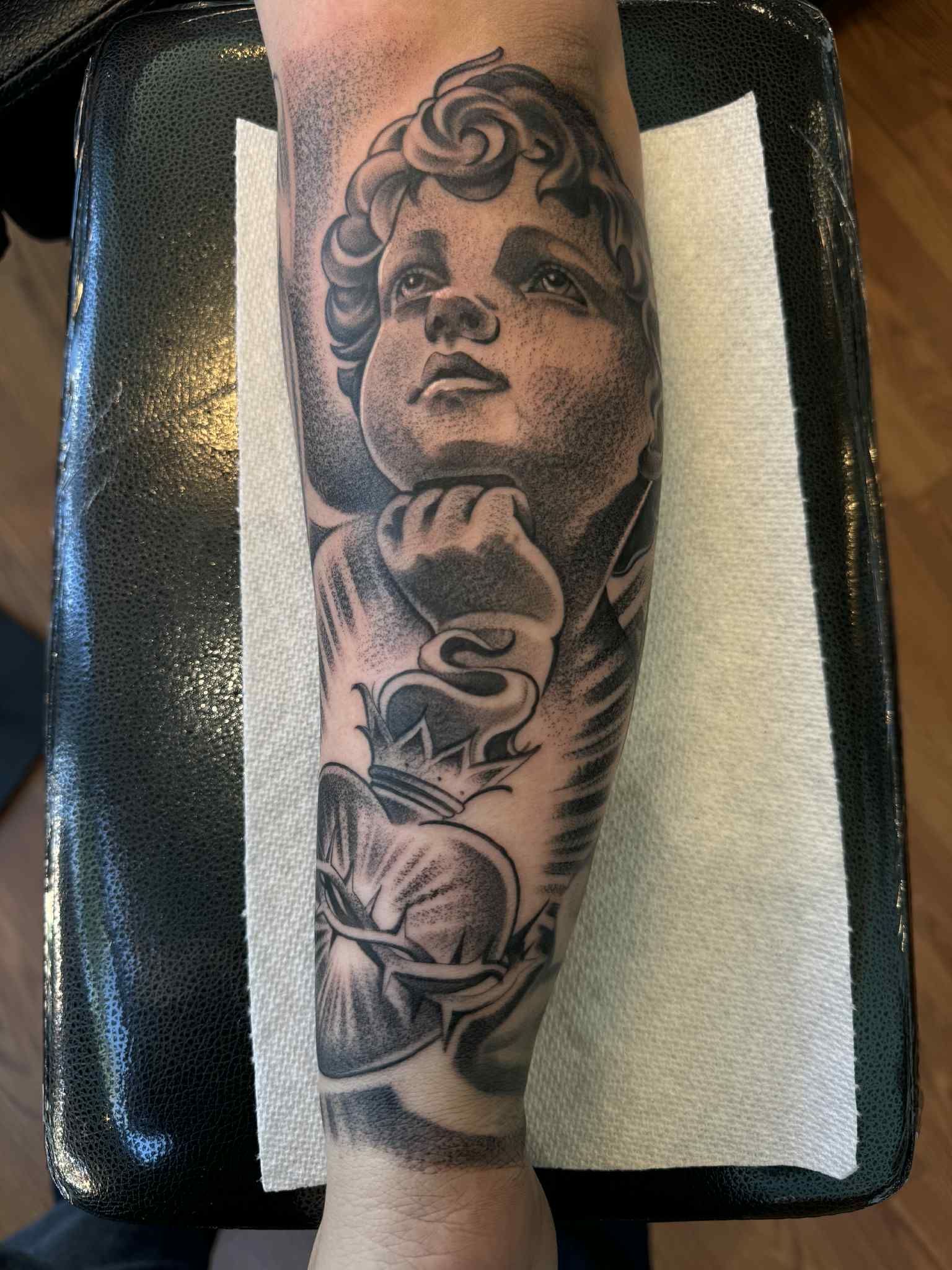 Black and white tattoo of cherub with a heart design on a forearm.