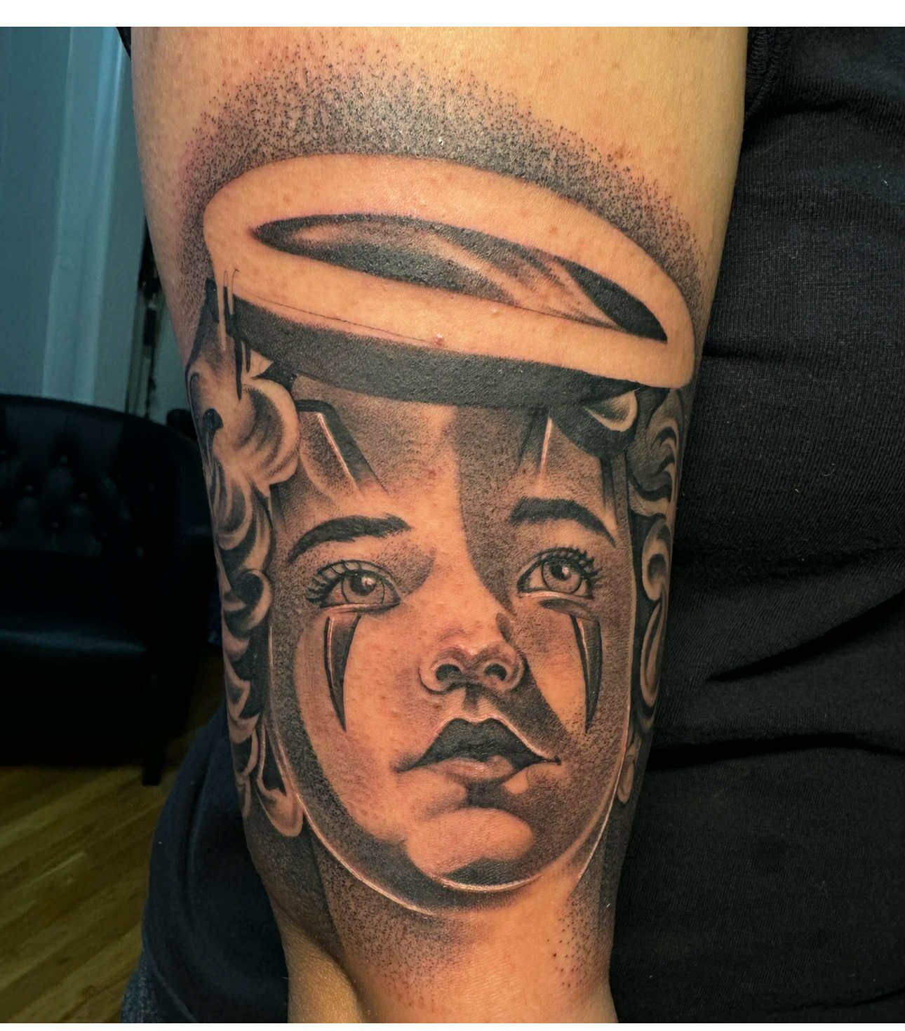Tattoo of a young face with a halo, shaded in black and gray, with tear-shaped markings.