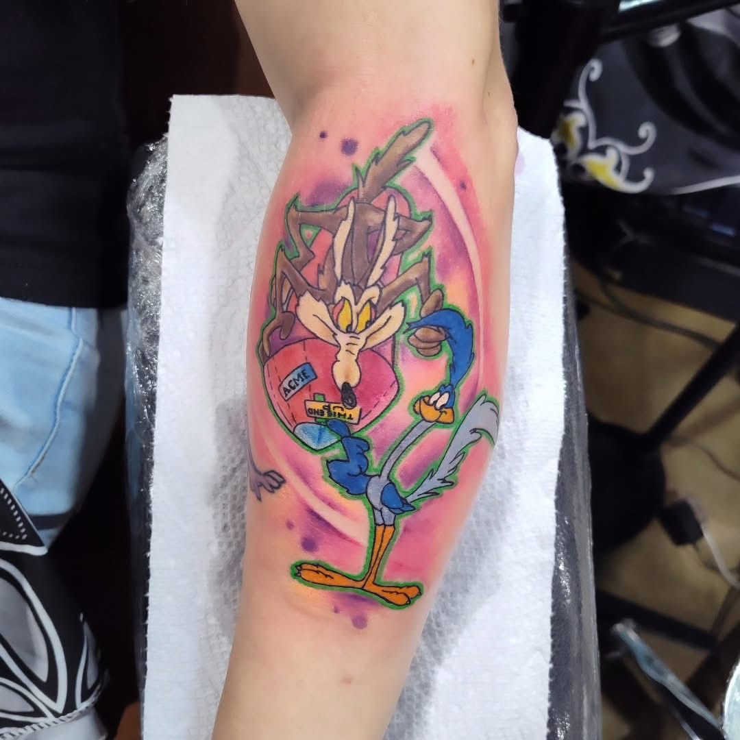 Tattoo of Wile E. Coyote and Road Runner with a pink and purple background on an arm.
