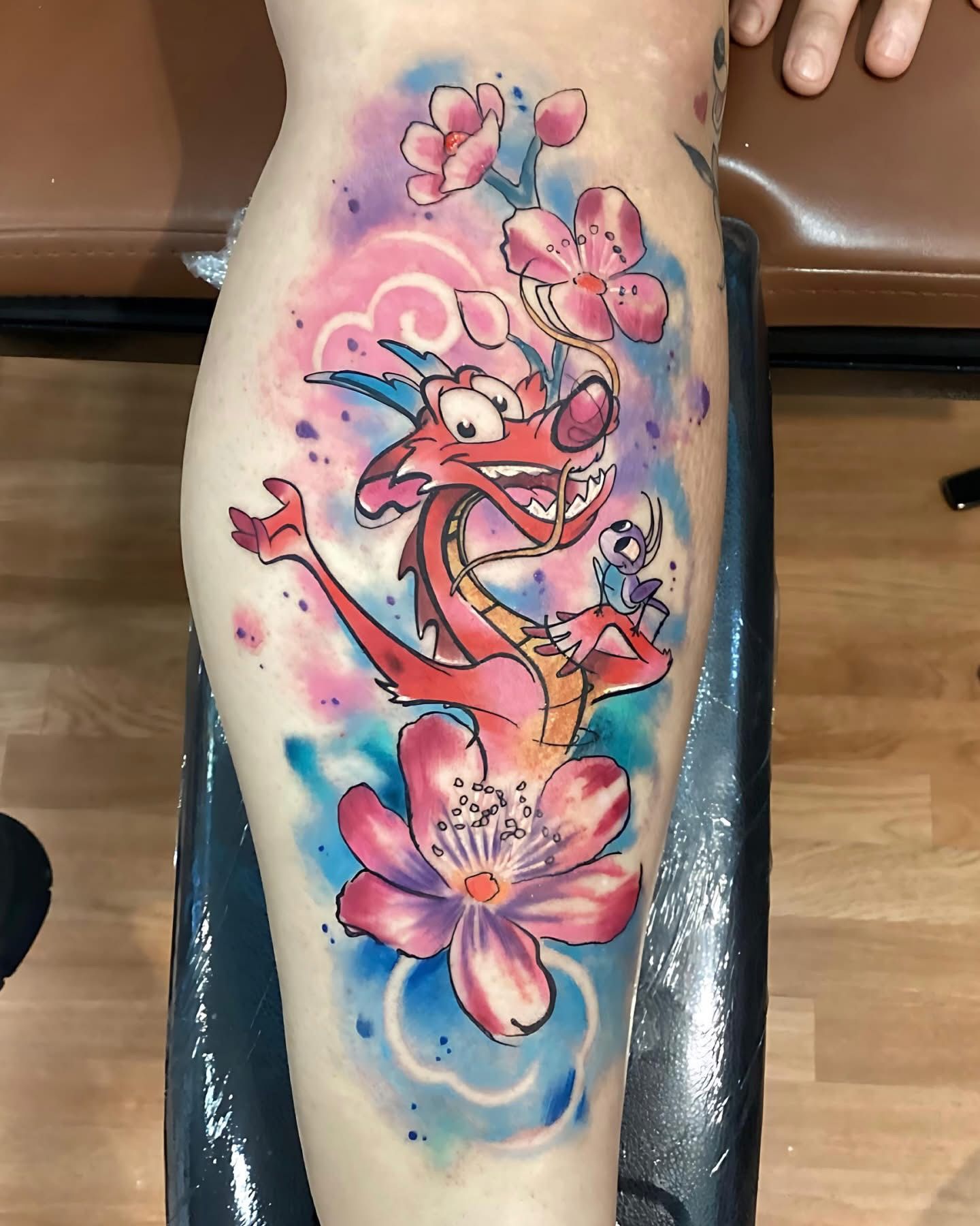 Mushu tattoo on a leg. Red dragon with pink blossoms and watercolor background.