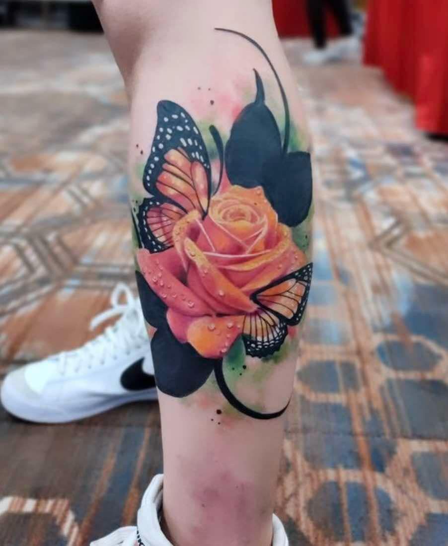 Tattoo of a rose and monarch butterflies on a leg; orange and black with watercolor effect.