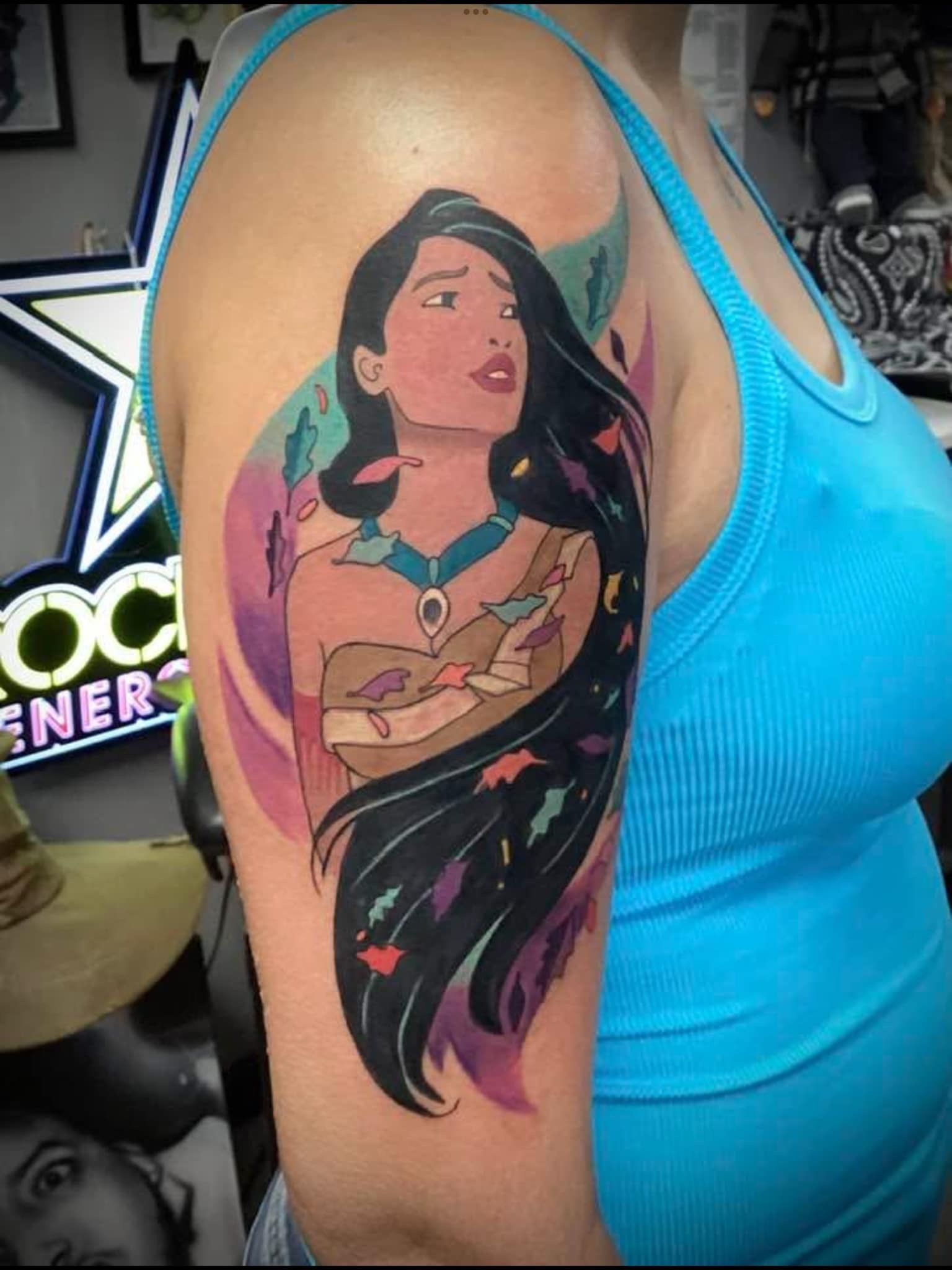 Pocahontas tattoo on a person's upper arm. Features her portrait with long black hair, surrounded by colorful leaves.
