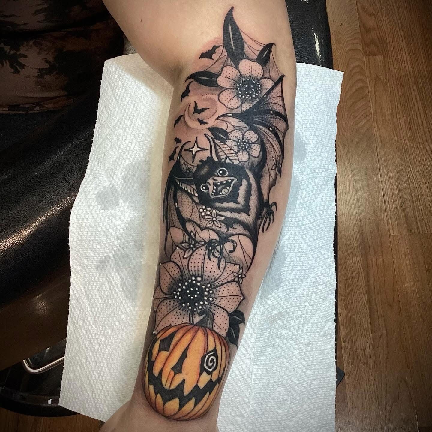 Tattoo of a Halloween-themed design on an arm; includes a pumpkin, flowers, a bat, and a spiderweb.