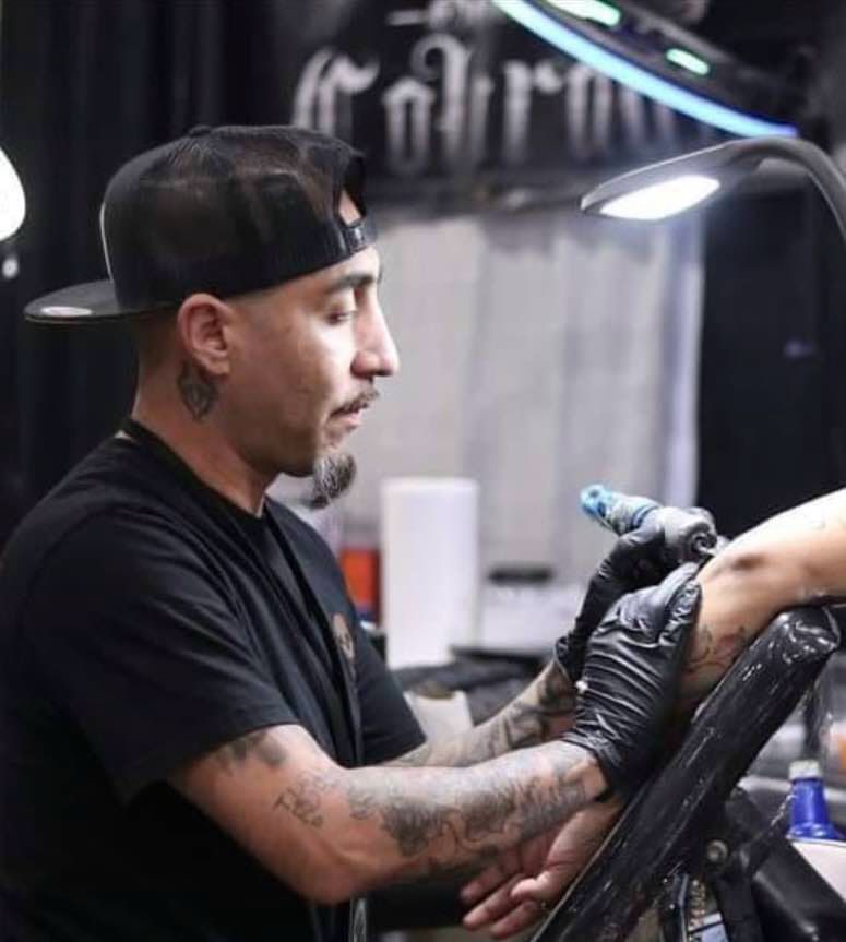 Tattoo artist with a black cap, wearing gloves, inking a person's arm. Studio setting with bright lamp.