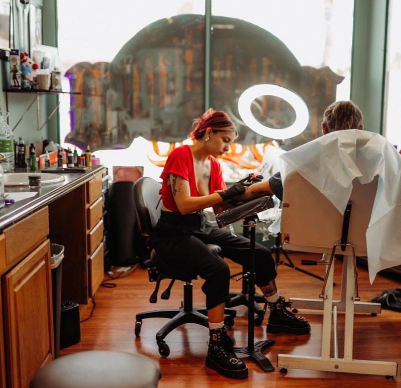 Tattoo artist in red shirt tattoos client's arm at a shop. Interior with wood cabinets and a round window.