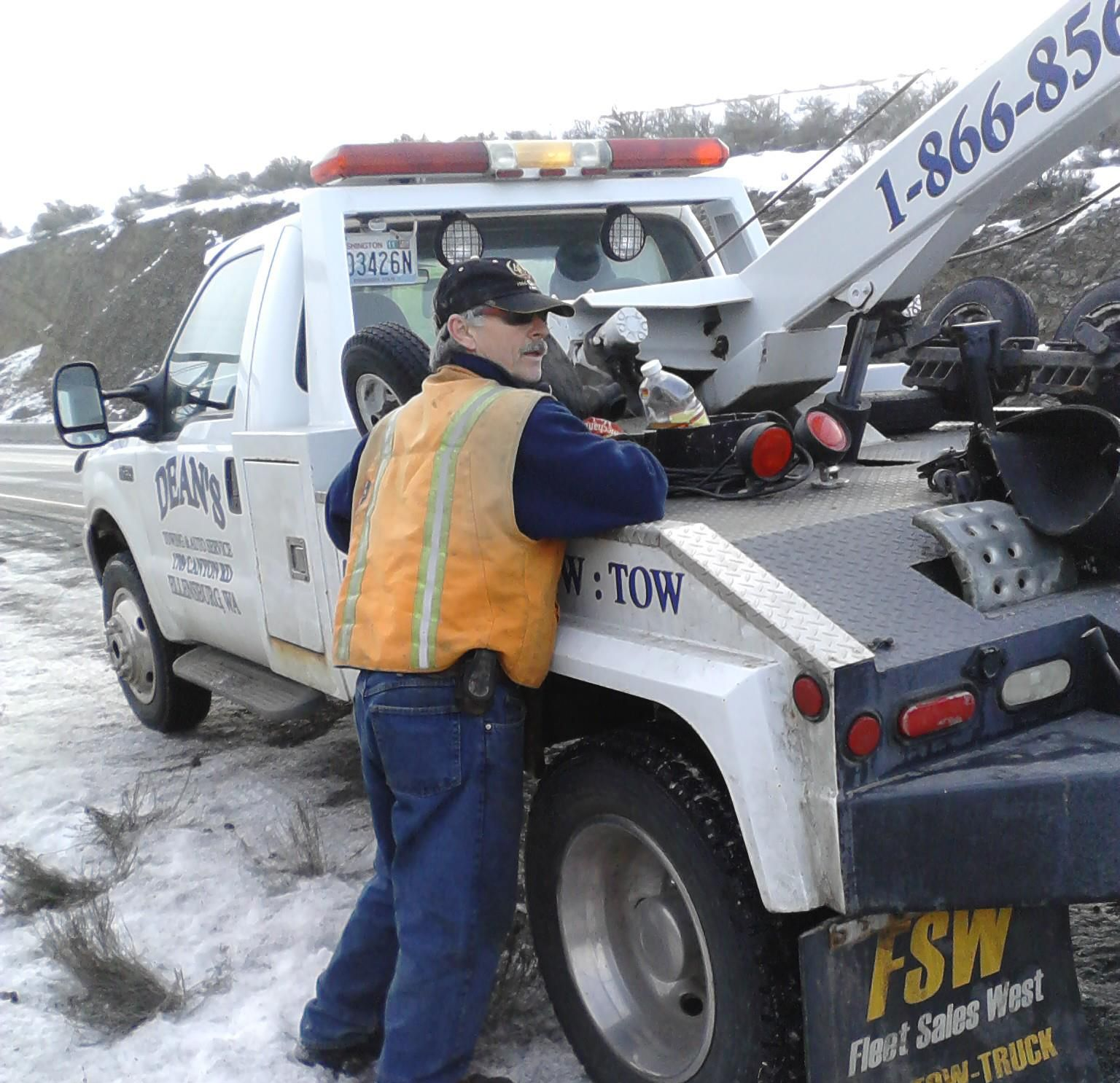 A worker in a safety vest leans against a white tow truck parked on a snowy roadside.