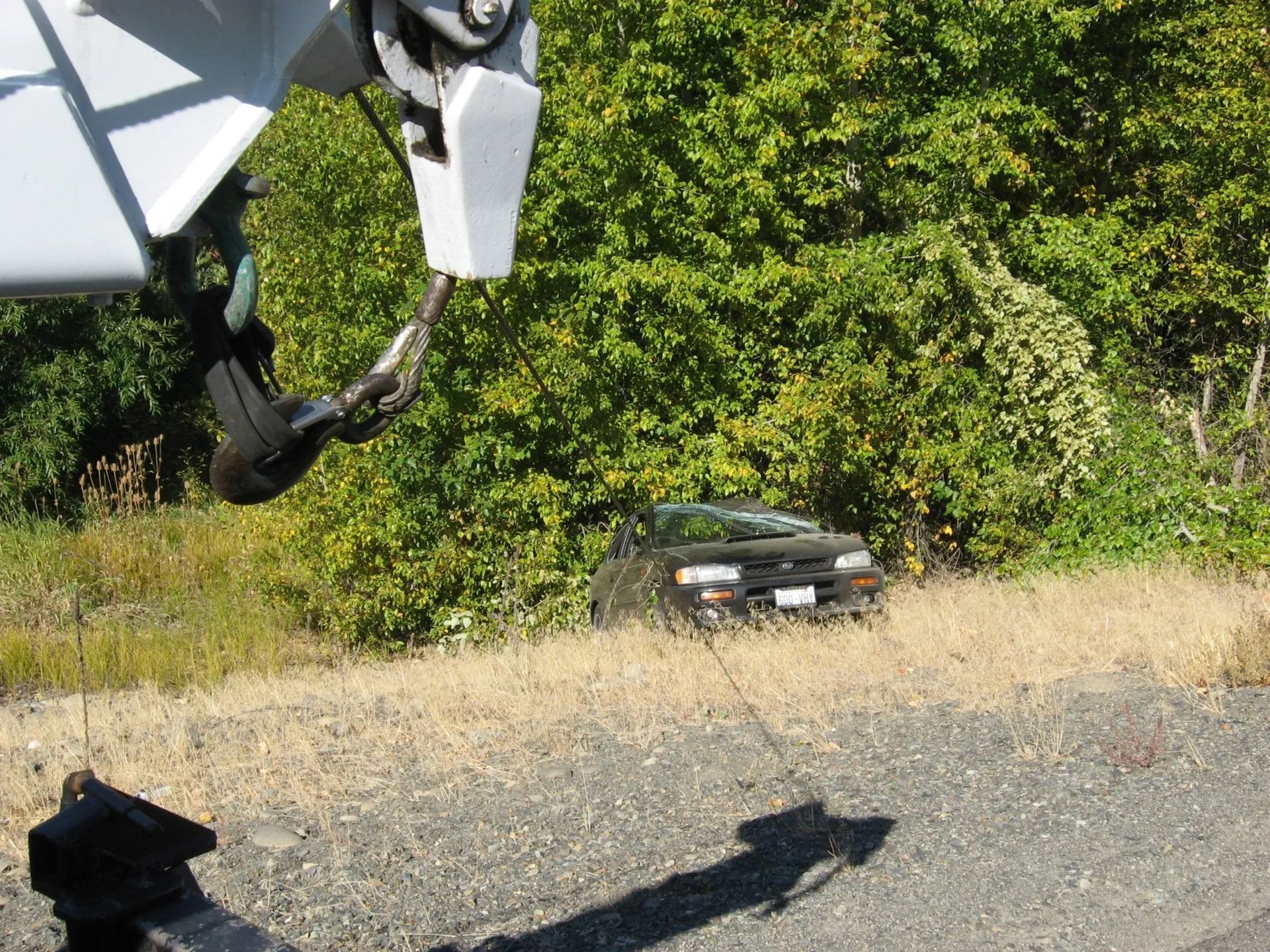 Black car on a gravel shoulder beside dense green bushes, with a vehicle shadow in the foreground.