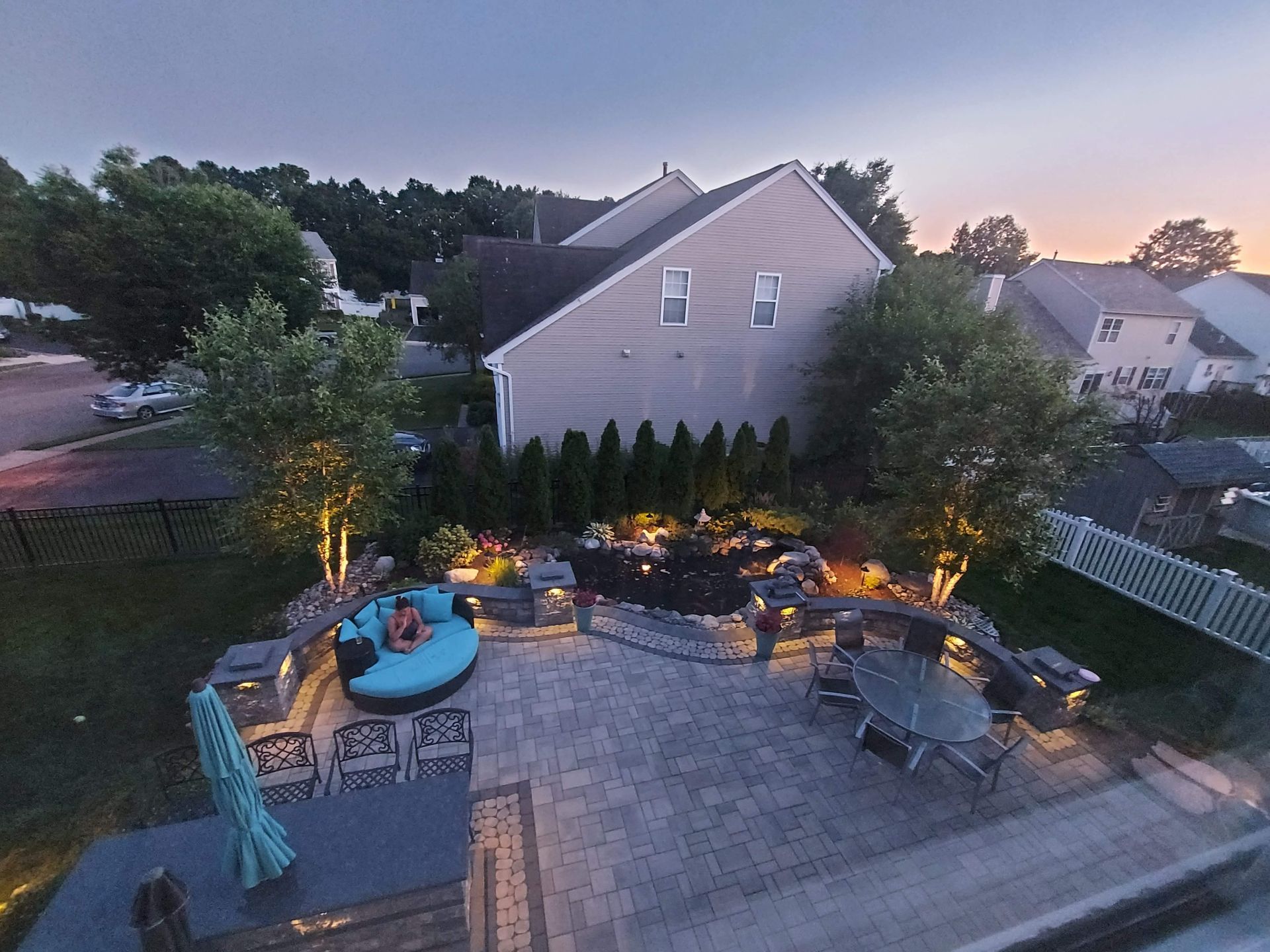 Three workers build a stone retaining wall in front of a residential house with a large pile of dirt and paved driveway. A dusk view of a paver patio featuring a round blue lounge, a dining set, built-in lighting, and a garden landscape.