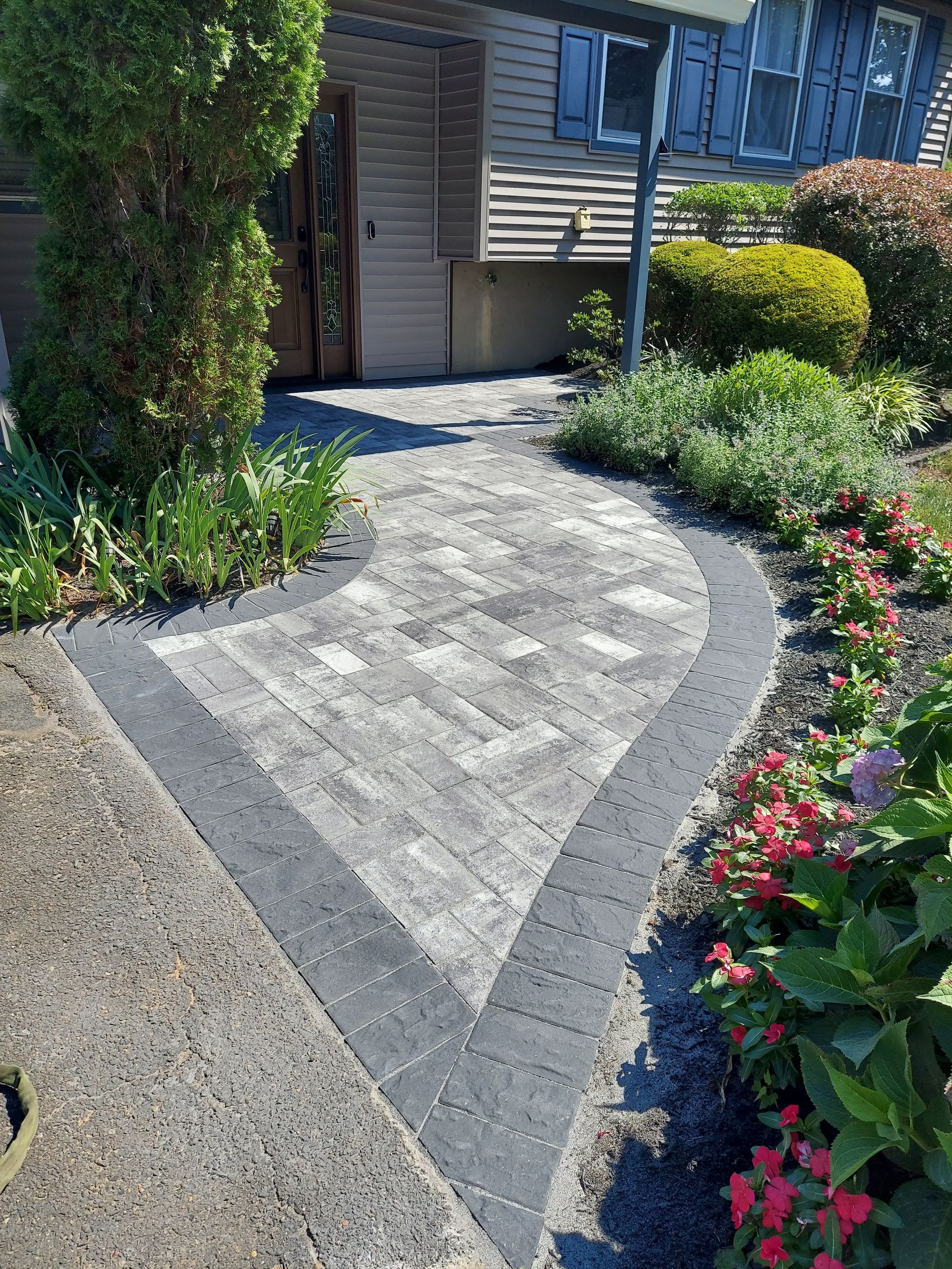A curved stone paver walkway with a dark border leads to the entrance of a house, surrounded by lush gardens and shrubs.
