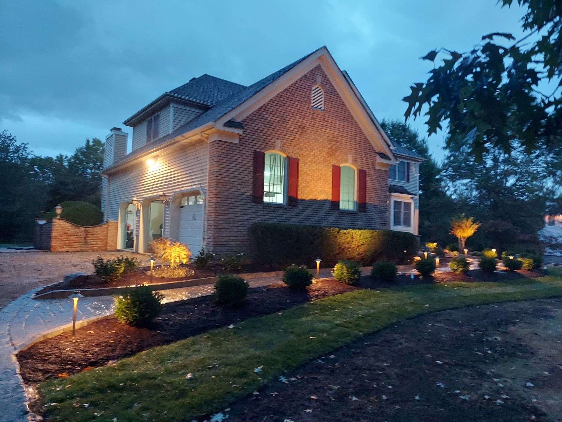 Two workers in white shirts stand in a large dirt trench outside a house, using tools to clear sand around a pipe. A two-story brick house at dusk, illuminated by glowing warm lights along the driveway and landscaped yard.