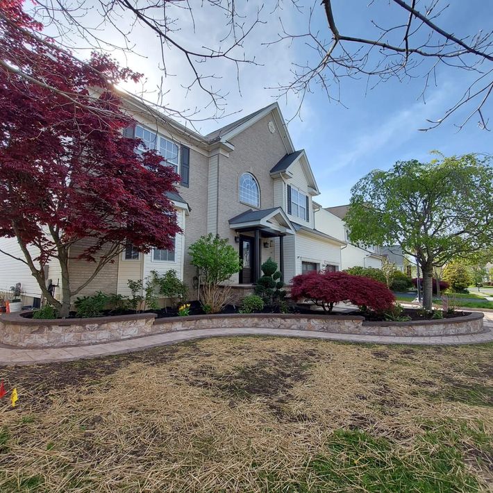 A two-story tan house with a stone retaining wall, front garden beds, a large red maple, and green trees under a blue sky. A two-story tan house with a stone retaining wall, front garden beds, a large red maple, and green trees under a blue sky.