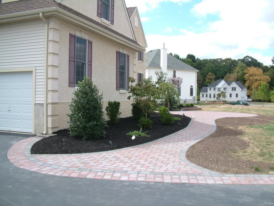 A tan house with a brick paver walkway leading to its front, surrounded by black mulch landscaping and other homes nearby. A tan house with a brick paver walkway leading to its front, surrounded by black mulch landscaping and other homes nearby.