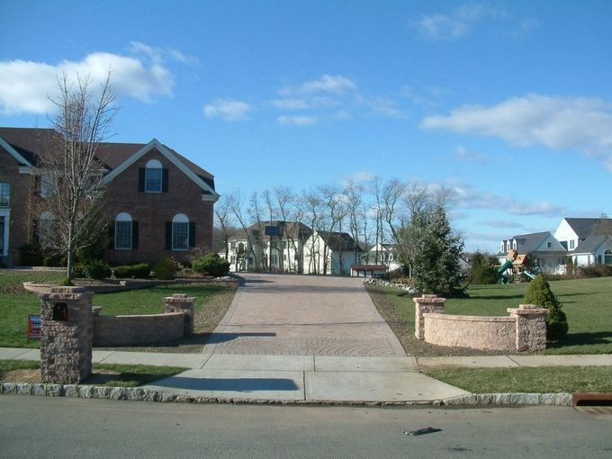 A long brick-paved driveway leads from a residential street toward a brick home under a clear blue sky. A long brick-paved driveway leads from a residential street toward a brick home under a clear blue sky.