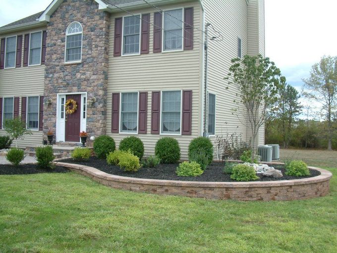 A two-story tan house with a stone facade entrance features a curved stone retaining wall surrounding a landscaped bed. A two-story tan house with a stone facade entrance features a curved stone retaining wall surrounding a landscaped bed.
