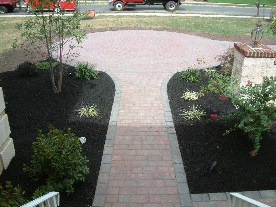 A brick pathway leads from a home's stairs to a circular patio, bordered by dark mulch and landscaping in a front yard. A brick pathway leads from a home's stairs to a circular patio, bordered by dark mulch and landscaping in a front yard.